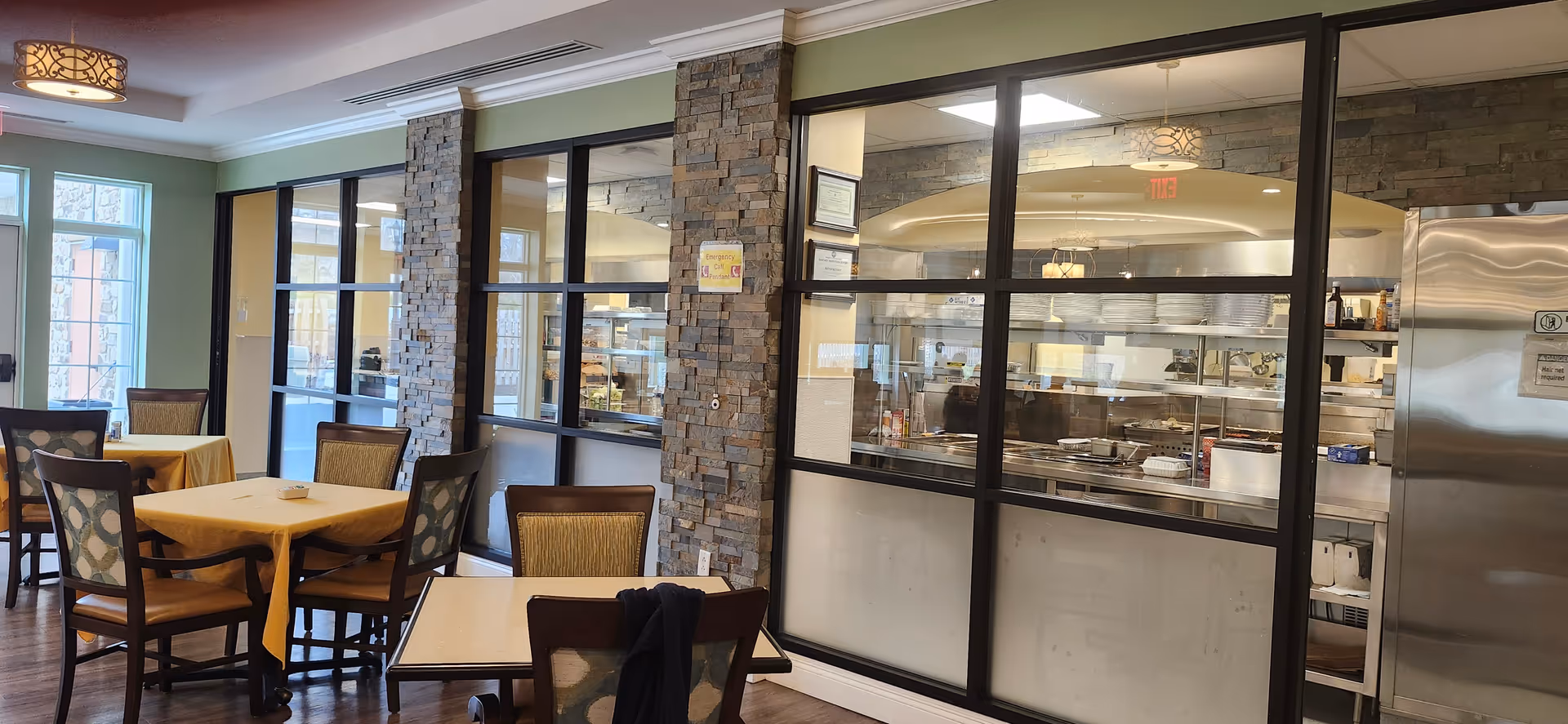 Dining area with tables covered in yellow tablecloths and chairs, adjacent to a kitchen visible through large glass windows framed with stone pillars. The kitchen has stainless steel appliances and shelves with plates. The room has green walls and wooden flooring.