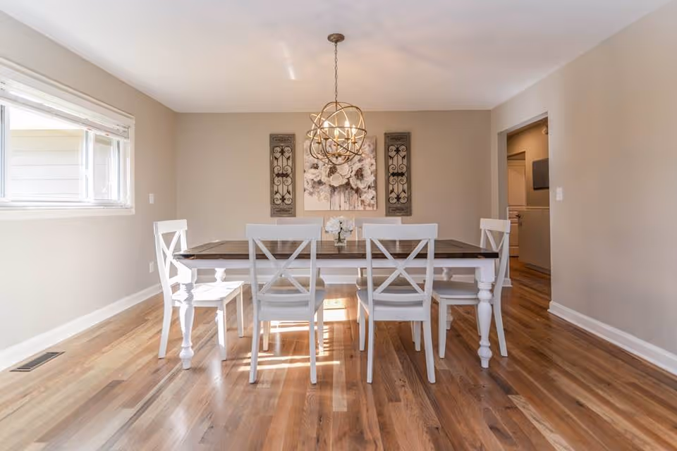 A bright dining room with a wooden floor, a rectangular wooden dining table with six white chairs around it, a modern spherical chandelier hanging above the table, and wall decorations including a floral painting and two decorative panels on either side.