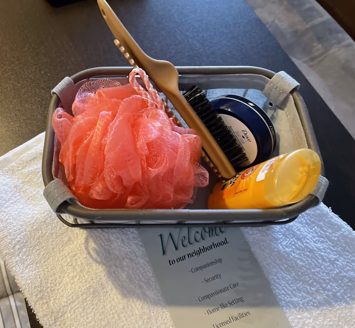 A small basket containing a pink bath pouf, a wooden brush, a container of Dove cream, and a yellow bottle of Arm & Hammer deodorant, placed on a white towel with a welcome note underneath.