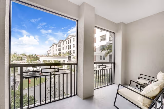 View from a covered balcony with two cushioned chairs overlooking a driveway and a multi-story residential building under a partly cloudy sky.