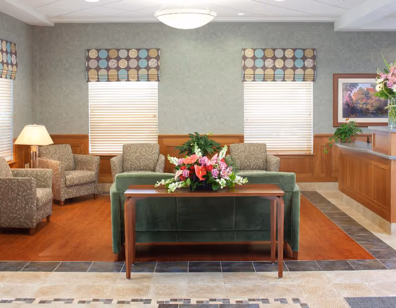 A well-lit sitting area in a senior living facility with a green sofa facing away from the camera, a wooden table with a colorful floral arrangement on it, three patterned armchairs, two windows with blinds and patterned valances, a lamp, a wooden reception desk with plants and a framed picture on the wall.