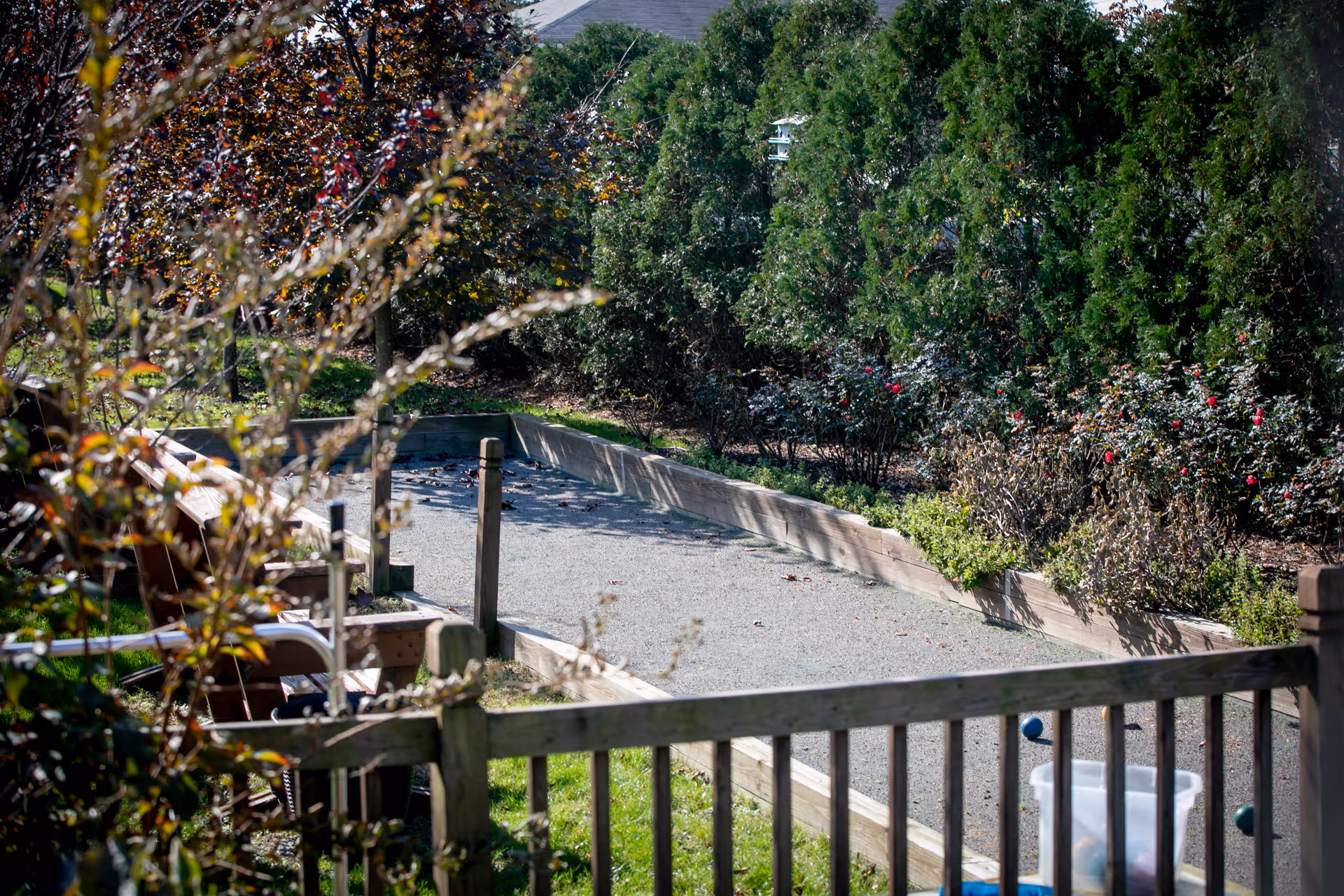 Outdoor bocce ball court surrounded by wooden fencing and greenery, including bushes and trees, with a few bocce balls and a plastic container visible near the court.