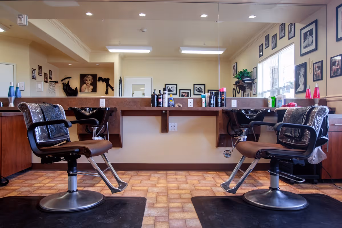 Interior view of a hair salon area with two brown salon chairs facing a large mirror. The countertop in front of the mirror holds various hair care products. The walls are decorated with framed black and white photographs and artwork. The floor has a patterned tile design.
