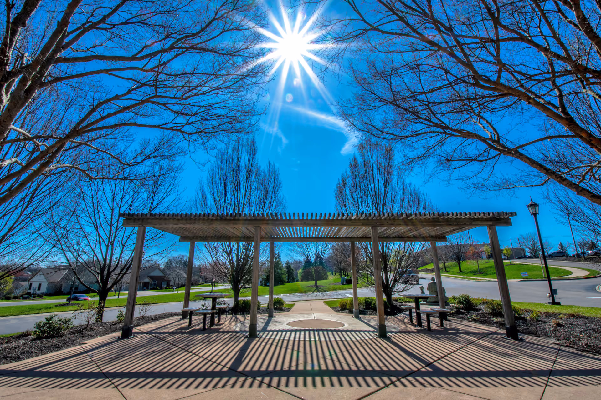 Outdoor seating area with a wooden pergola casting striped shadows on the concrete ground, surrounded by leafless trees under a bright blue sky with the sun shining directly above.