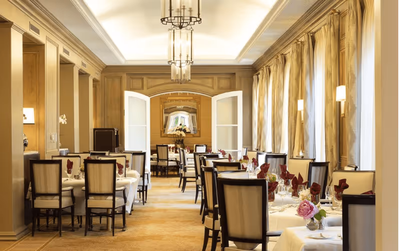 Elegant dining room with multiple tables set with white tablecloths, glassware, and red napkins. The room features beige walls, large windows with beige curtains, wall sconces, and two hanging chandeliers. There is a mirror on the far wall reflecting part of the room.