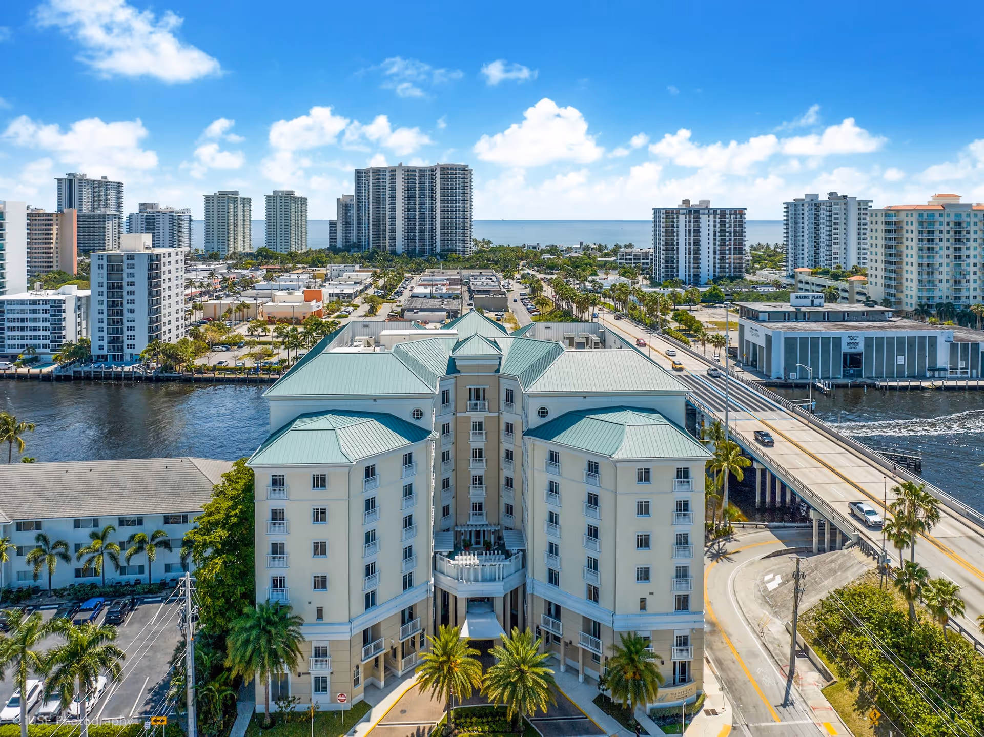 Aerial view of a large multi-story building with a green roof, surrounded by palm trees and roads, with a bridge crossing over water nearby. In the background, there are multiple high-rise buildings and a clear blue sky with some clouds.