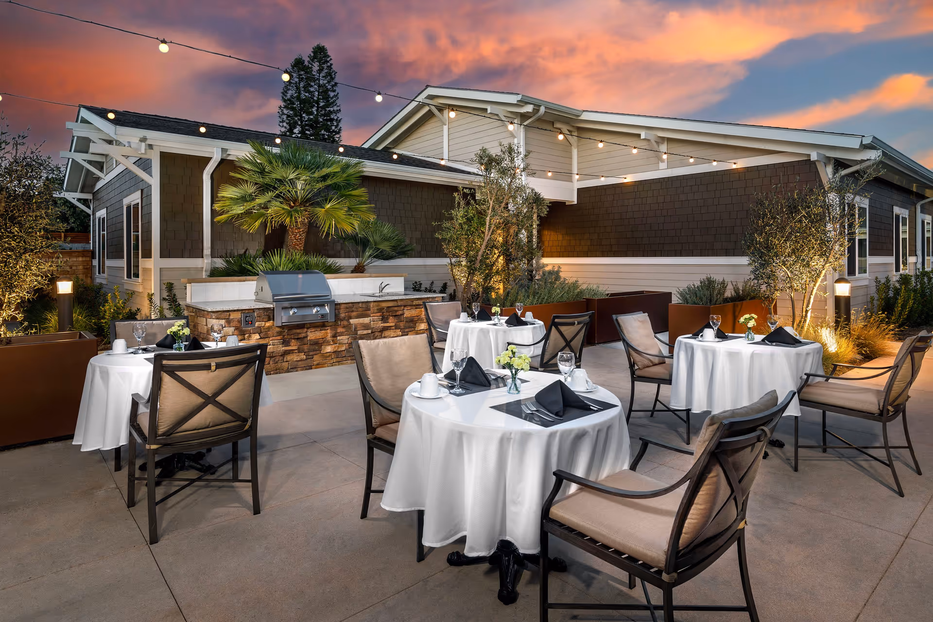 Outdoor patio dining area at sunset with round tables set with white tablecloths and chairs in front of a single-story building.