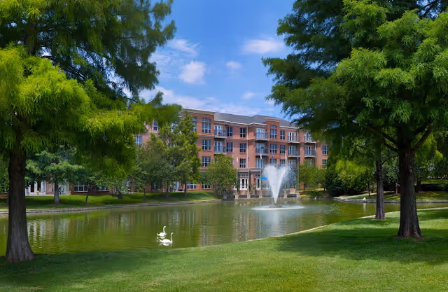 A serene outdoor scene featuring a pond with a fountain in the center, surrounded by green grass and tall trees. In the background, there is a multi-story brick building with balconies. Two white swans are swimming on the pond under a partly cloudy blue sky.