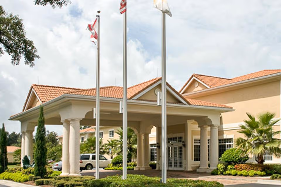 Covered entrance of a Mediterranean-style building with columns, three flagpoles, and manicured landscaping.