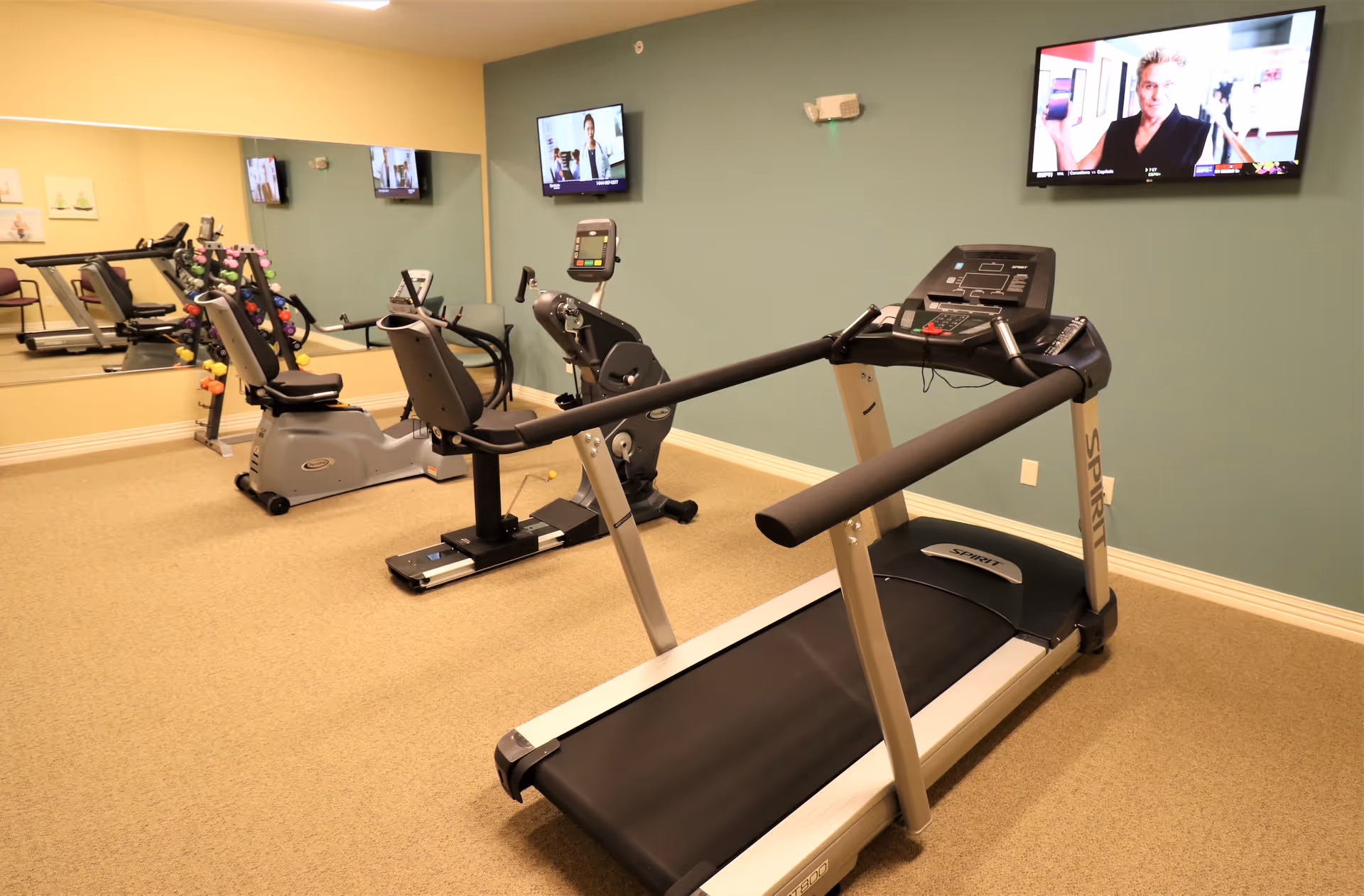 A small fitness room with exercise equipment including a treadmill, a recumbent bike, and a stationary bike. There is a rack of colorful dumbbells against the wall with a large mirror reflecting the room. Two flat-screen TVs are mounted on the walls, one showing a man holding a phone and the other showing a woman in a white coat.