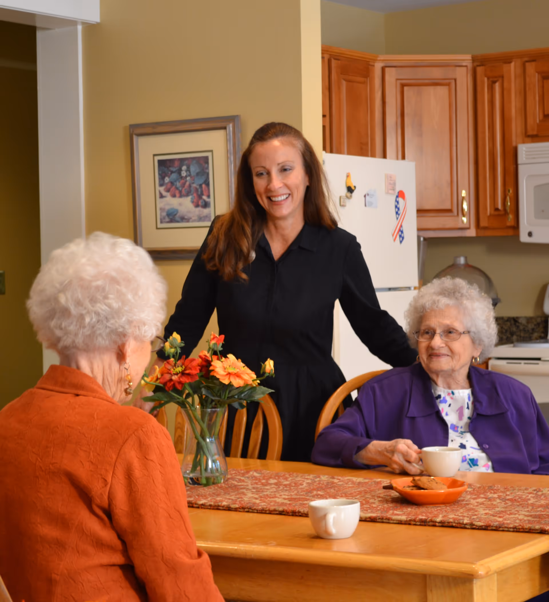 Two elderly women sitting at a wooden dining table with a vase of flowers and cups, while a younger woman stands behind them smiling in a kitchen setting with wooden cabinets and a refrigerator.