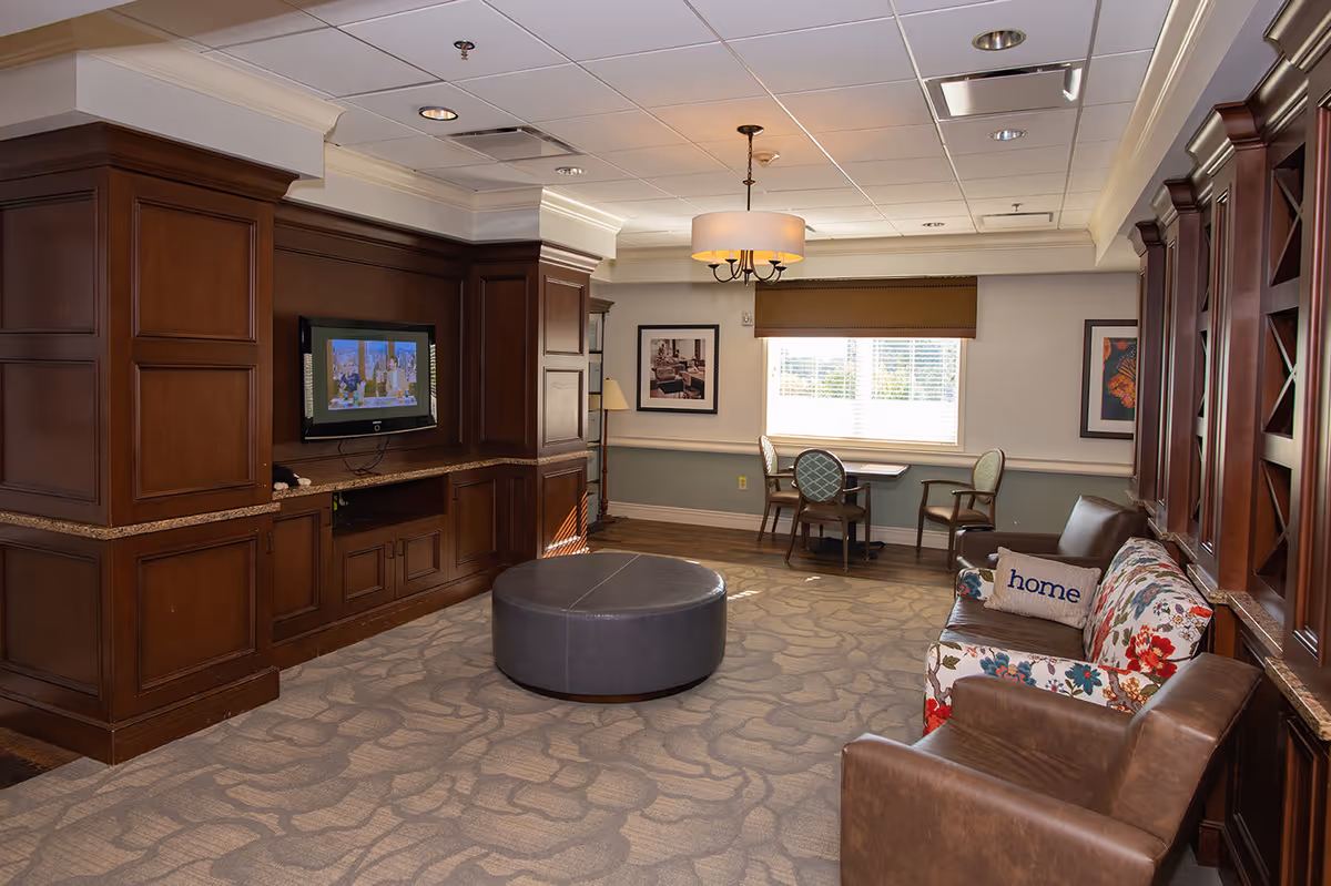 A cozy senior living common room with wood cabinetry, a wall-mounted TV, a round ottoman, and seating including a floral sofa and armchairs near a windowed table.