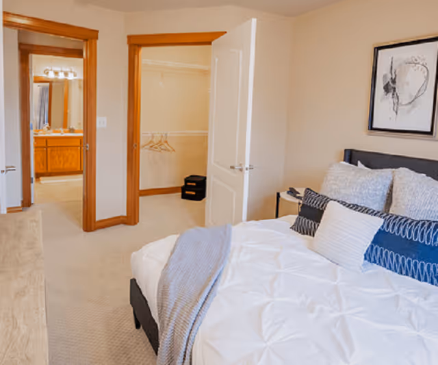 A bedroom with a neatly made bed featuring white and blue pillows and a gray throw blanket. The room has beige walls and carpet, a wooden dresser, and a framed abstract artwork on the wall. Two open doors reveal a walk-in closet with hangers and a bathroom with a wooden vanity and mirror.