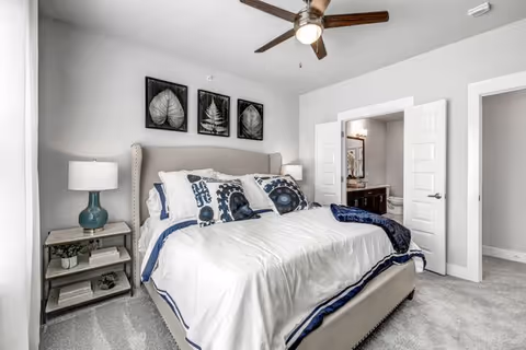 A bright bedroom with a large bed featuring white and blue bedding, two bedside tables with lamps, three framed leaf artworks above the headboard, a ceiling fan with light, and an open doorway leading to a bathroom.