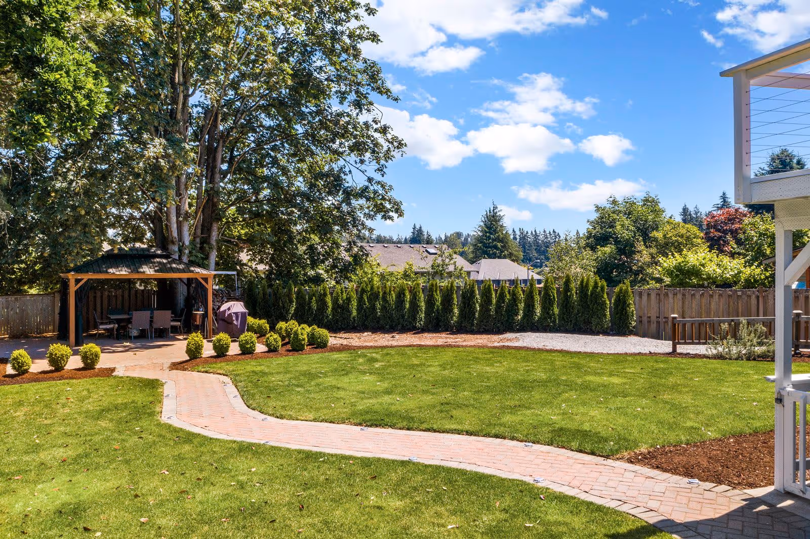 A well-maintained outdoor garden area with a green lawn, a curved brick pathway, and a wooden gazebo with outdoor furniture underneath. The garden is surrounded by tall trees, shrubs, and a wooden fence, with a clear blue sky and some clouds overhead.