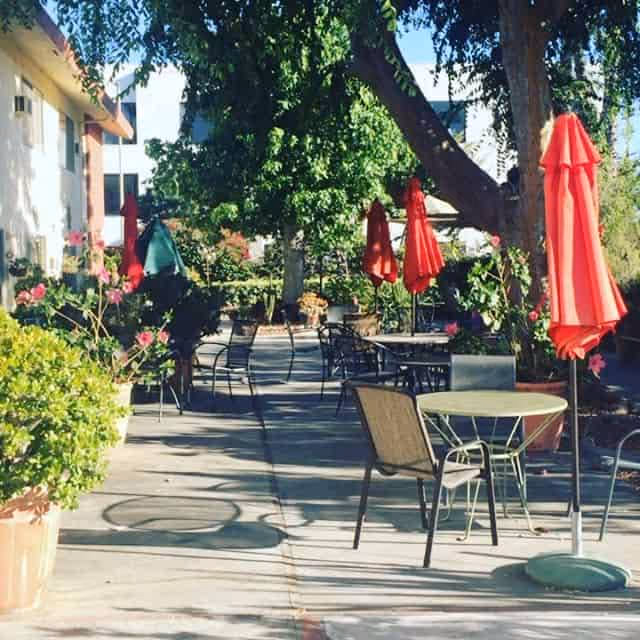 Outdoor patio area with several round tables and chairs, some tables have red umbrellas. The patio is shaded by large trees and surrounded by plants and flowers.