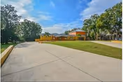 Wide concrete driveway leading to a yellow building with a red roof, surrounded by green grass and trees under a blue sky with scattered clouds.