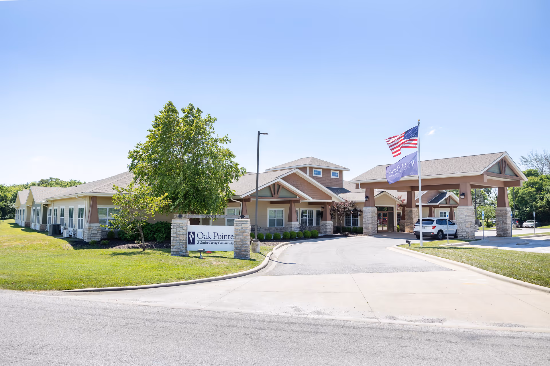 Exterior front view of Oak Pointe of Neosho senior living community building with a driveway, an American flag and a community flag on flagpoles, green lawn, and clear blue sky.