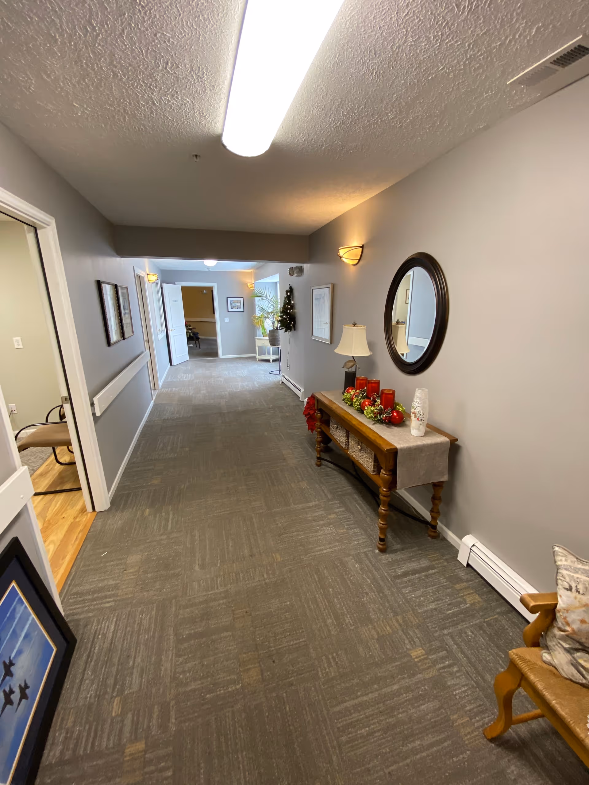 A well-lit hallway in a senior living facility with gray walls and carpeted floor. On the right side, there is a wooden console table decorated with red candles, a lamp, and holiday greenery, beneath a round mirror. Further down the hallway, there is a small decorated Christmas tree and a chair near a window. On the left side, there is an open door leading to a room with wooden flooring and a chair. The hallway has ceiling lights and wall sconces providing illumination.