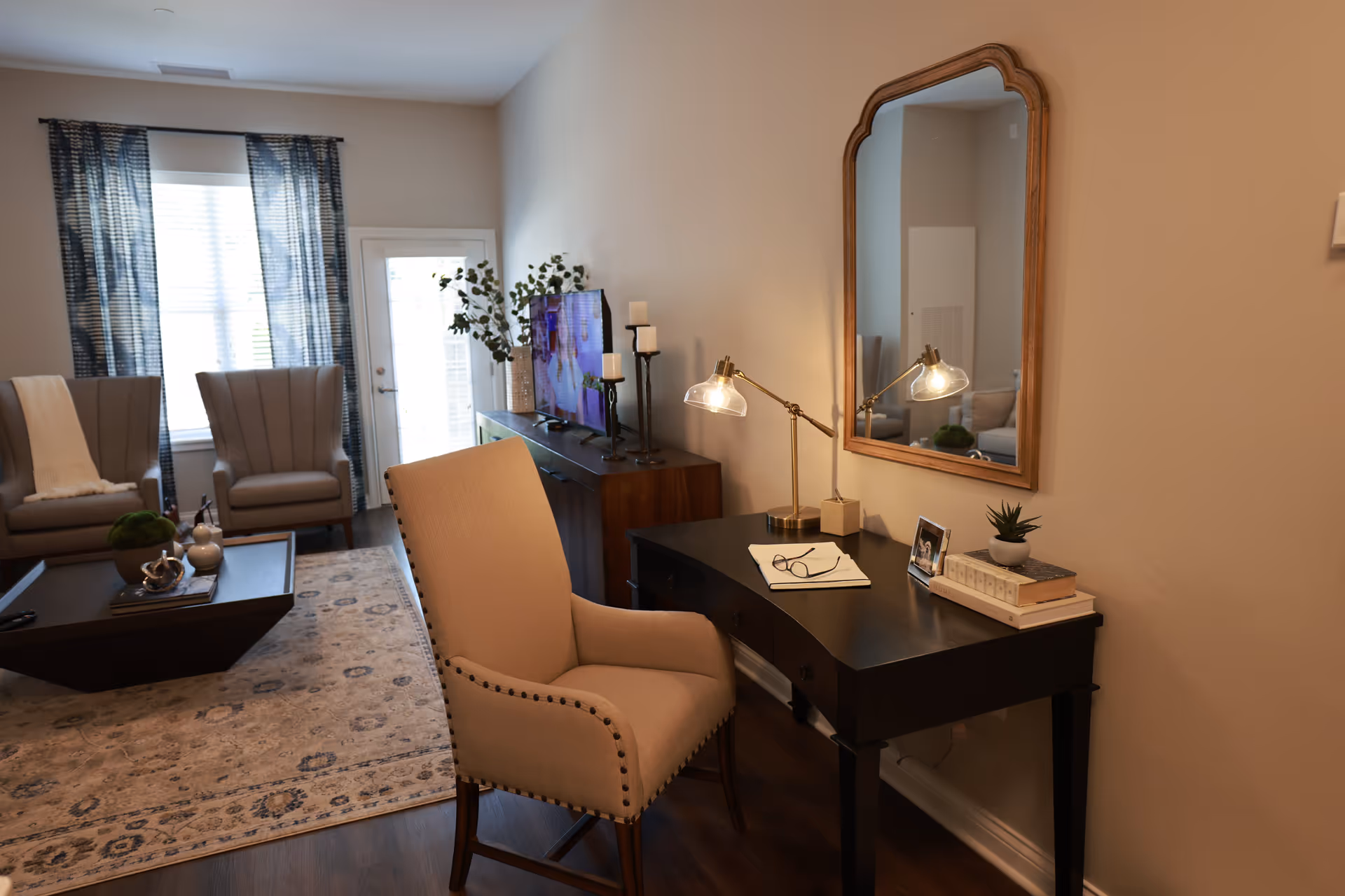 A cozy living room area with two beige armchairs near a window with blue patterned curtains. A dark wooden coffee table with decorative items sits on a patterned rug. A black desk with a beige upholstered chair is against the wall, featuring a table lamp, books, a small plant, a photo frame, and a pair of glasses on an open notebook. A large mirror with a wooden frame hangs above the desk. A TV and decorative candles are on a wooden console table along the wall.