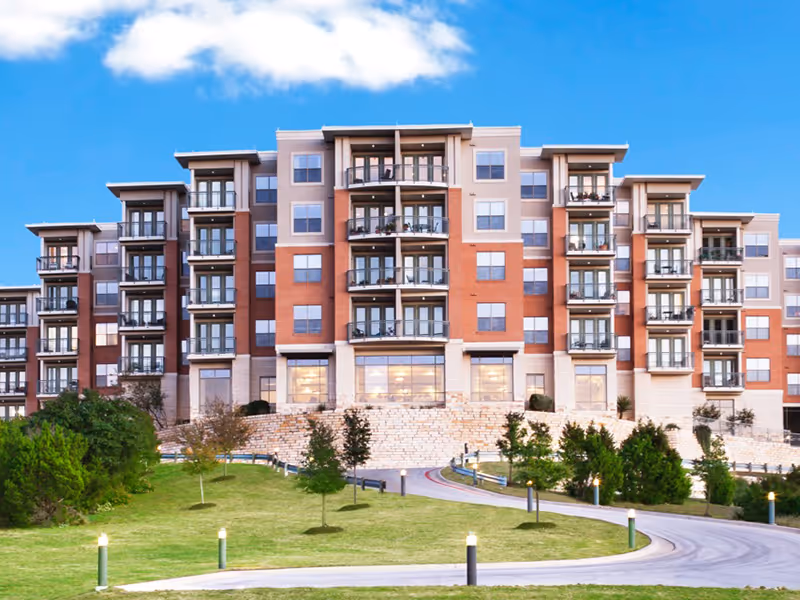 A large multi-story residential building with balconies, surrounded by a landscaped lawn and trees under a blue sky with some clouds.