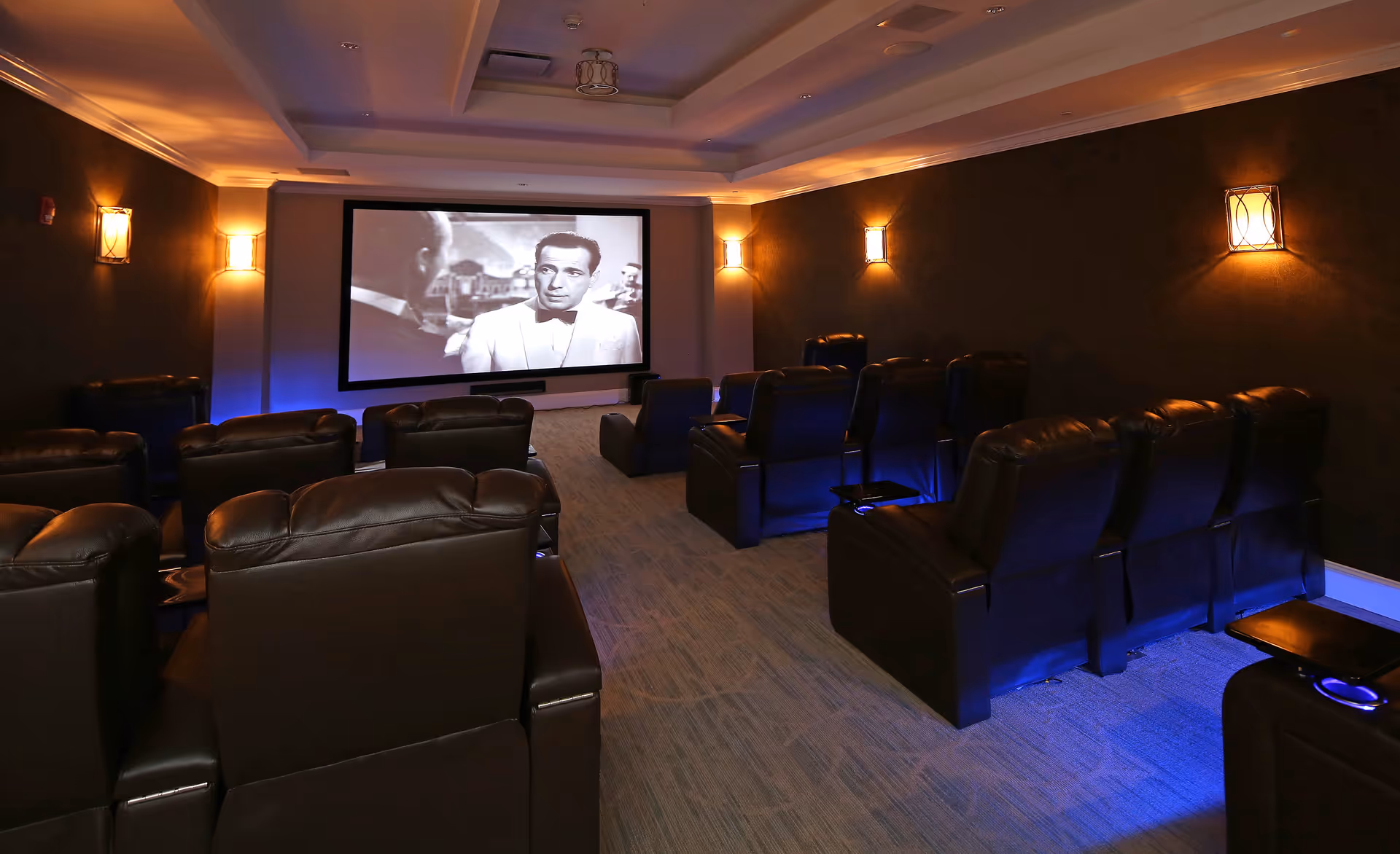 A cozy movie theater room with multiple rows of dark leather recliner chairs facing a large screen showing a black and white movie. The room has dim wall sconces providing warm lighting and blue accent lights near the floor.