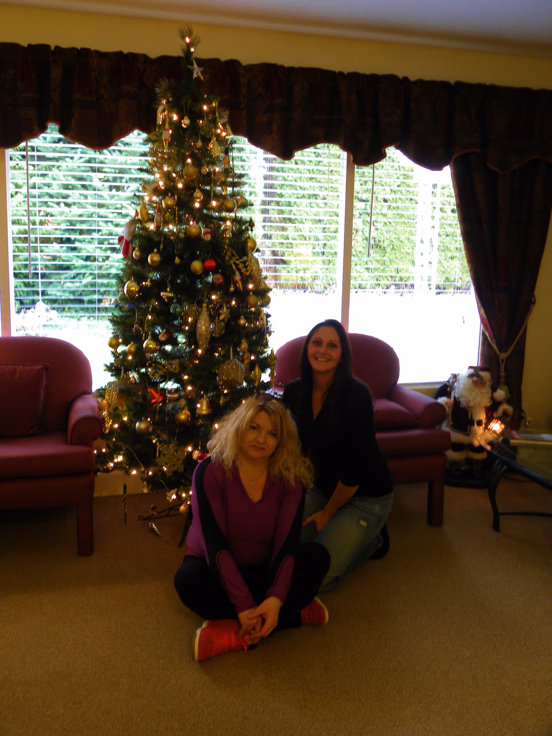 Two women sitting on the carpeted floor in front of a decorated Christmas tree with lights in a living room. Behind them are two red armchairs, large windows with blinds, and dark curtains. A Santa Claus decoration is visible near a glass coffee table on the right side.