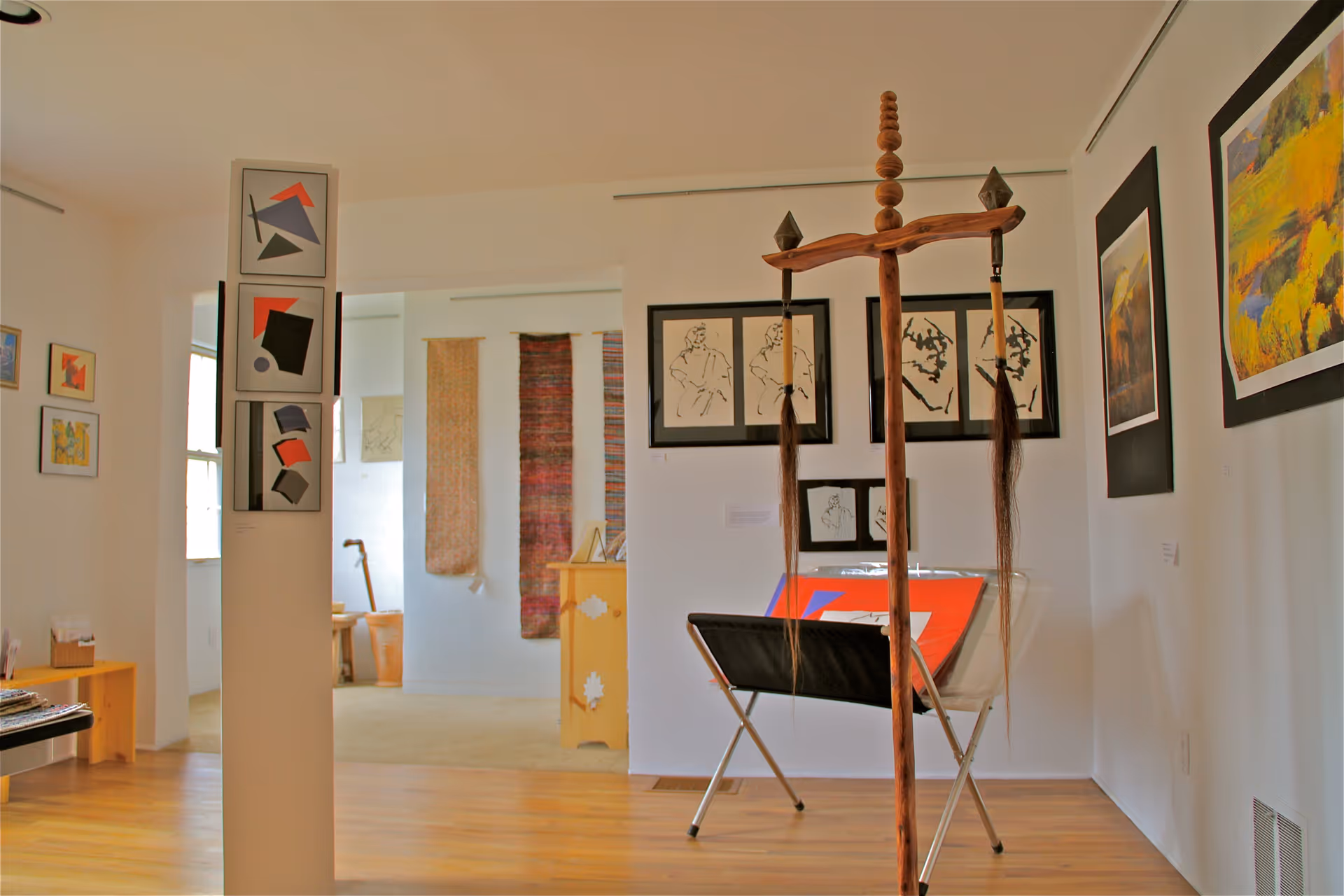 Interior view of an art gallery or exhibition space with various framed artworks on the walls, a wooden sculpture with hanging elements in the foreground, and textile art pieces hanging in the background. The room has wooden flooring and white walls.