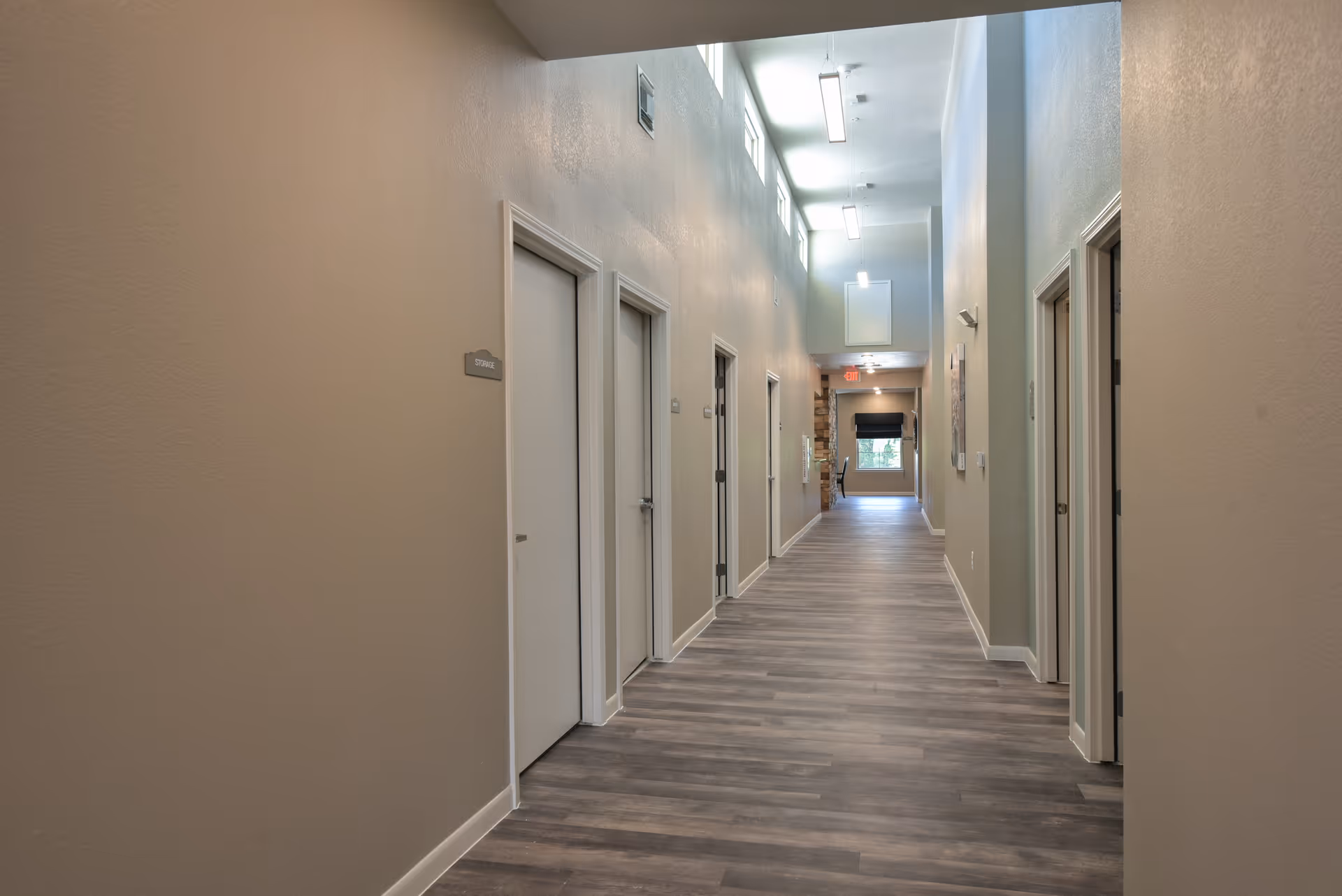 A long, well-lit hallway in a senior living facility with multiple closed doors on both sides. The floor has wood-like vinyl planks, and the walls are painted beige. Small windows near the ceiling allow natural light to enter, and fluorescent lights hang from the ceiling. A sign on one door reads 'STORAGE'.