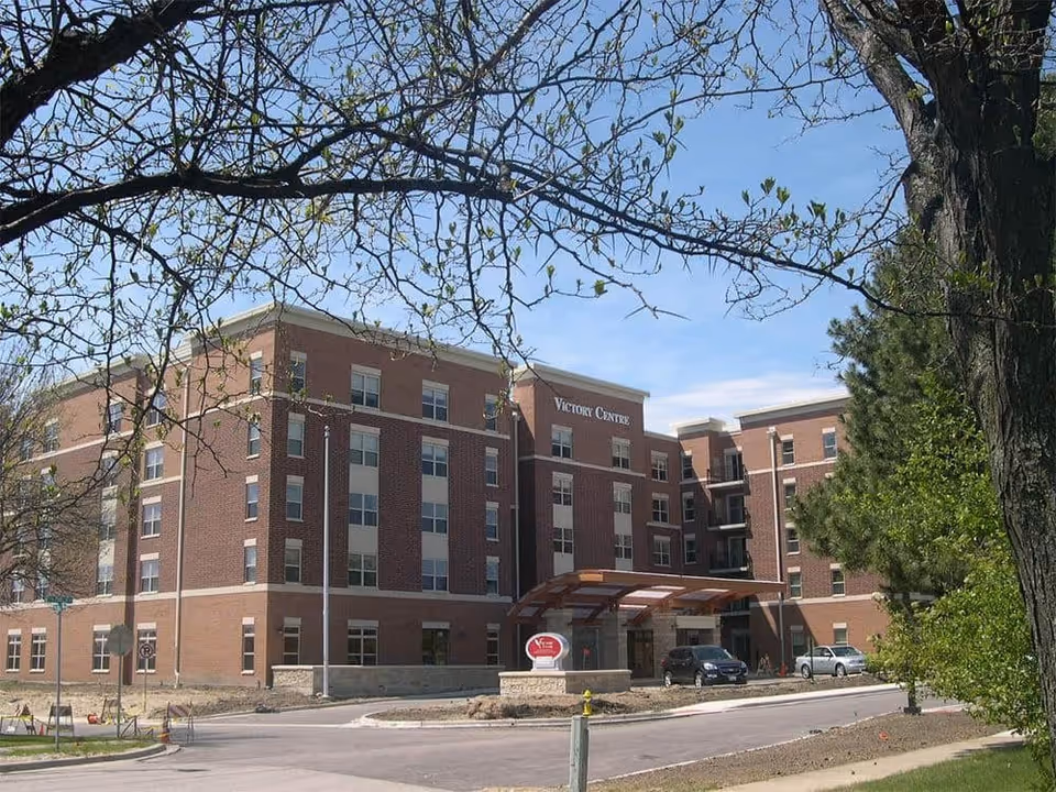 Exterior view of a multi-story brick building named Victory Centre, surrounded by trees and a driveway with parked cars in front.