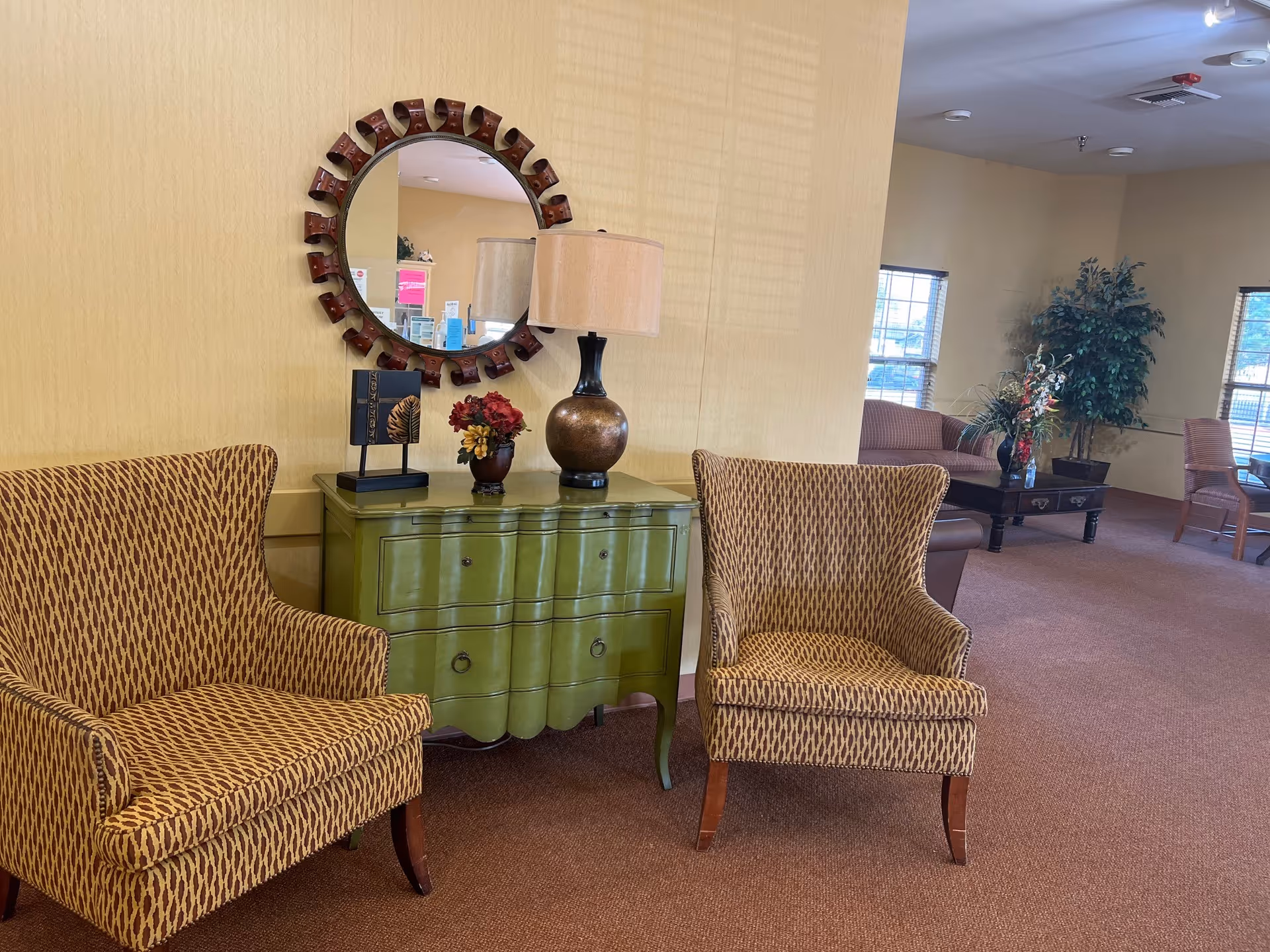 A cozy interior lounge area with two patterned armchairs flanking a green chest of drawers. On top of the chest are a decorative lamp, a small flower arrangement, and a decorative book. A round mirror with a unique frame hangs on the yellow wall above the chest. In the background, there are additional seating areas with sofas, chairs, a coffee table, and potted plants near windows.