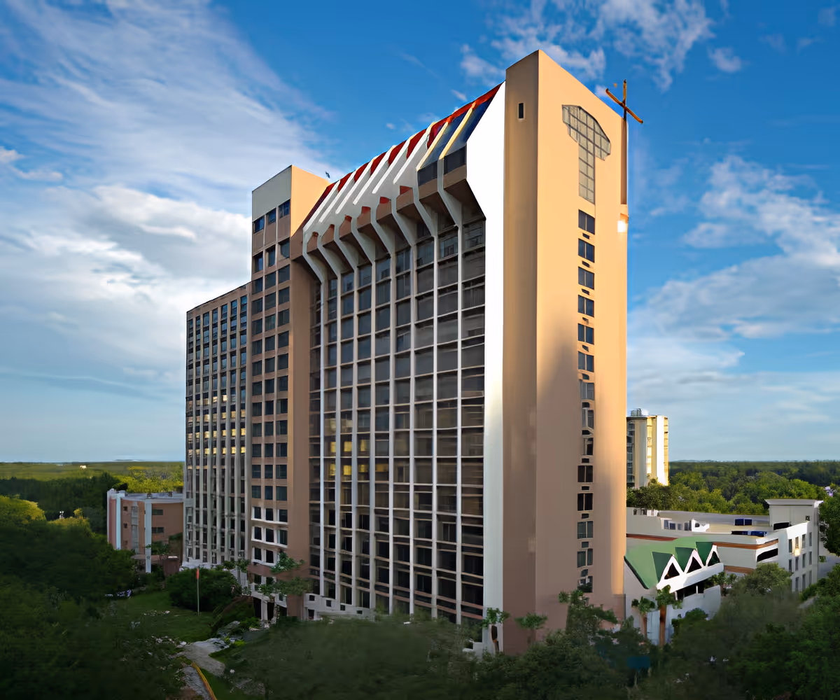 Tall multi-story building with large vertical windows and a cross mounted on the corner, surrounded by greenery under a partly cloudy blue sky.