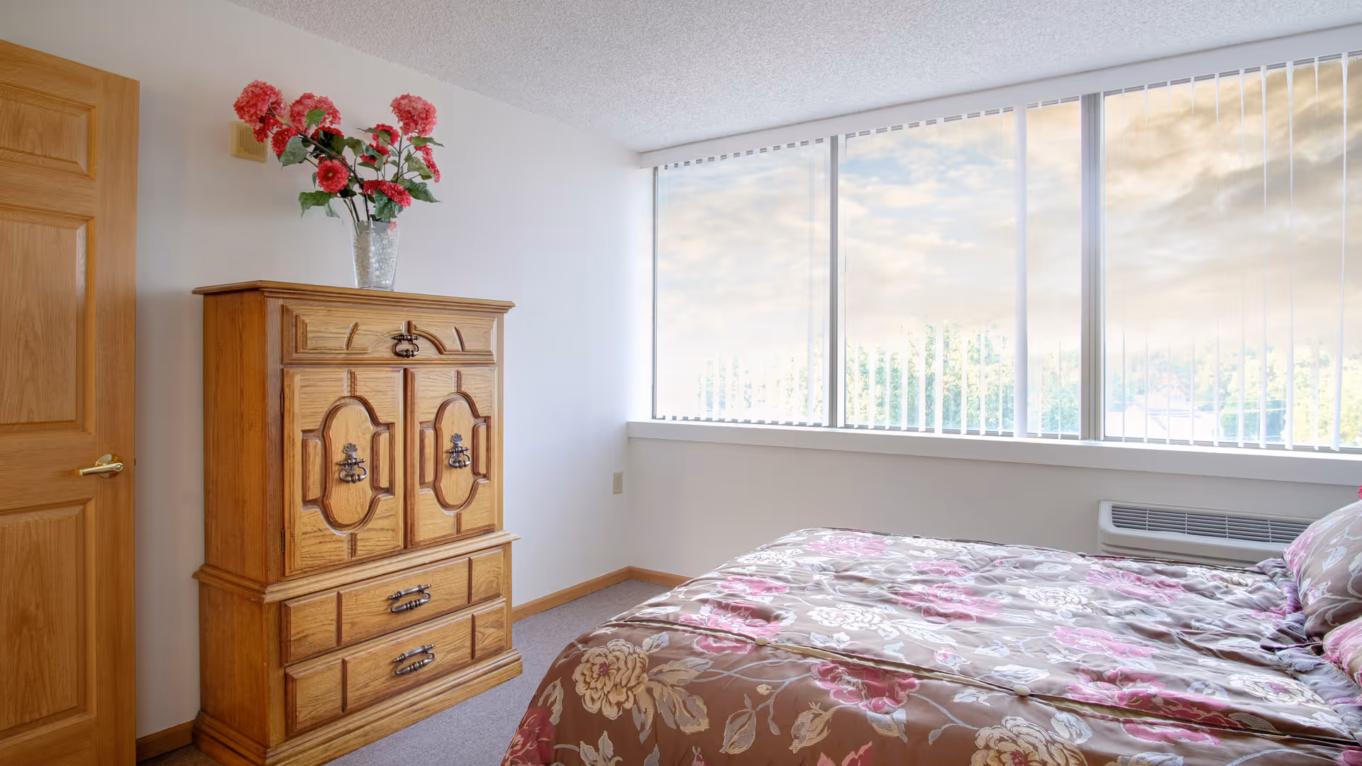 A bright bedroom with a large window covered by vertical blinds, a wooden cabinet with decorative handles and a vase of red flowers on top, and a bed with a floral-patterned comforter in shades of brown, pink, and white.