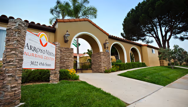 Exterior view of Arroyo Vista Nursing Center showing a beige building with arched entryways, stone pillars, a well-maintained lawn, and a sign displaying the facility name and address at 3022 45th Street.