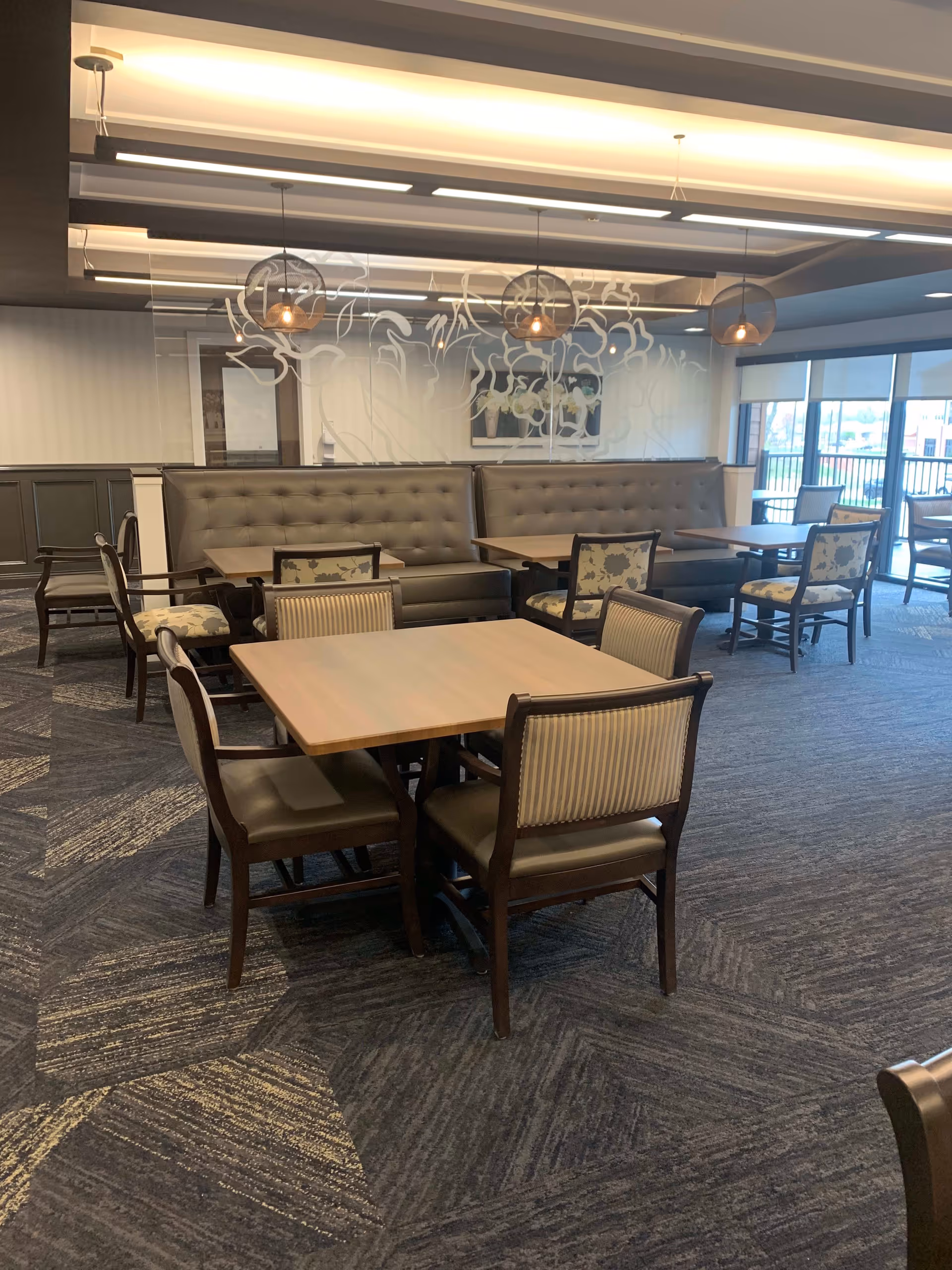 A dining area with multiple wooden tables and chairs. The chairs have cushioned seats and backs with different fabric patterns. There are two long tufted leather benches against a glass wall with floral designs etched on it. Pendant lights hang from the ceiling, and large windows on the right side let in natural light.