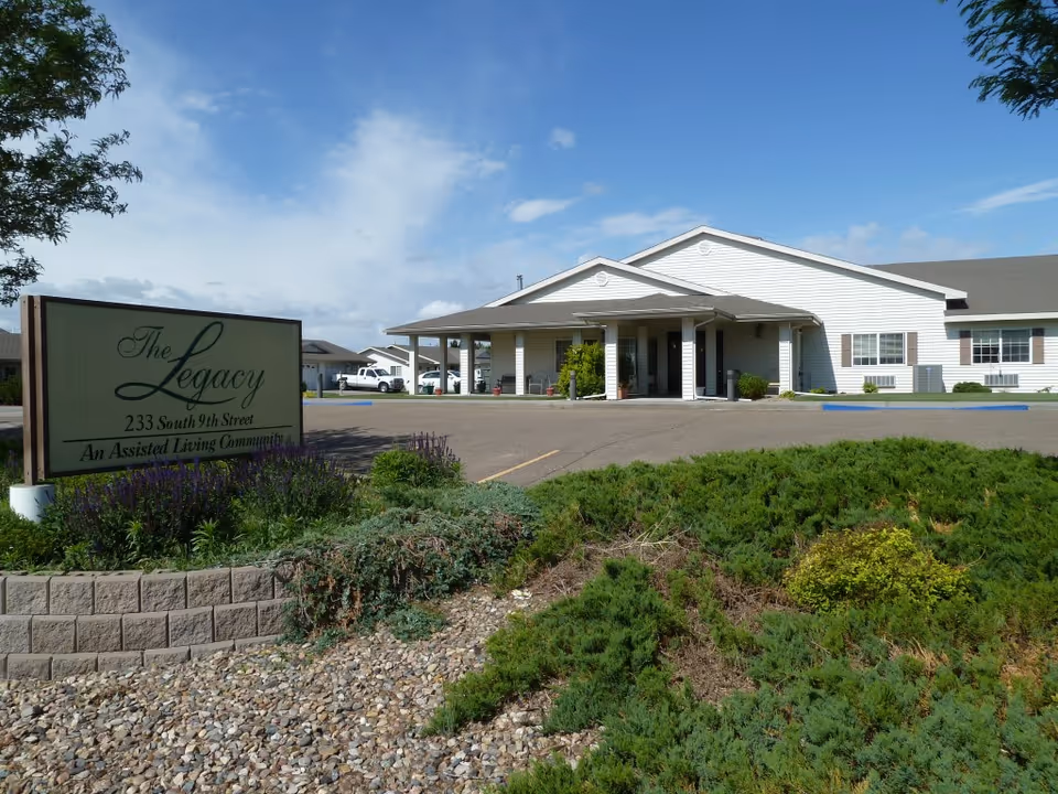 Front exterior of The Legacy assisted living facility with entrance canopy, parking area, and a landscaped sign.