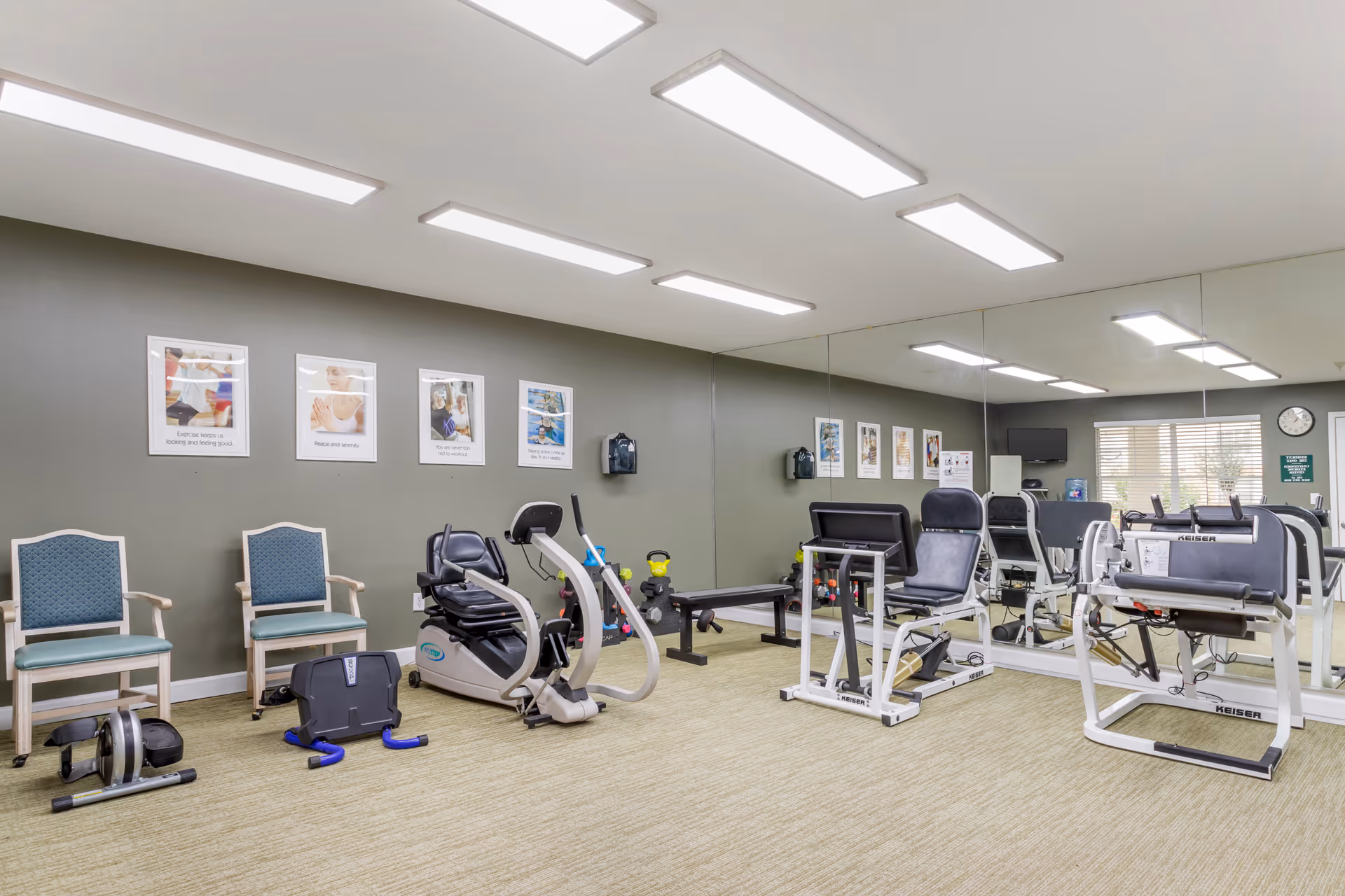 A well-lit exercise room with various fitness equipment including a recumbent bike, leg press machines, and weights. Two chairs are placed against a gray wall adorned with motivational exercise posters. A large mirror covers one wall, reflecting the room and equipment. A clock and a small TV are visible near a window with blinds.