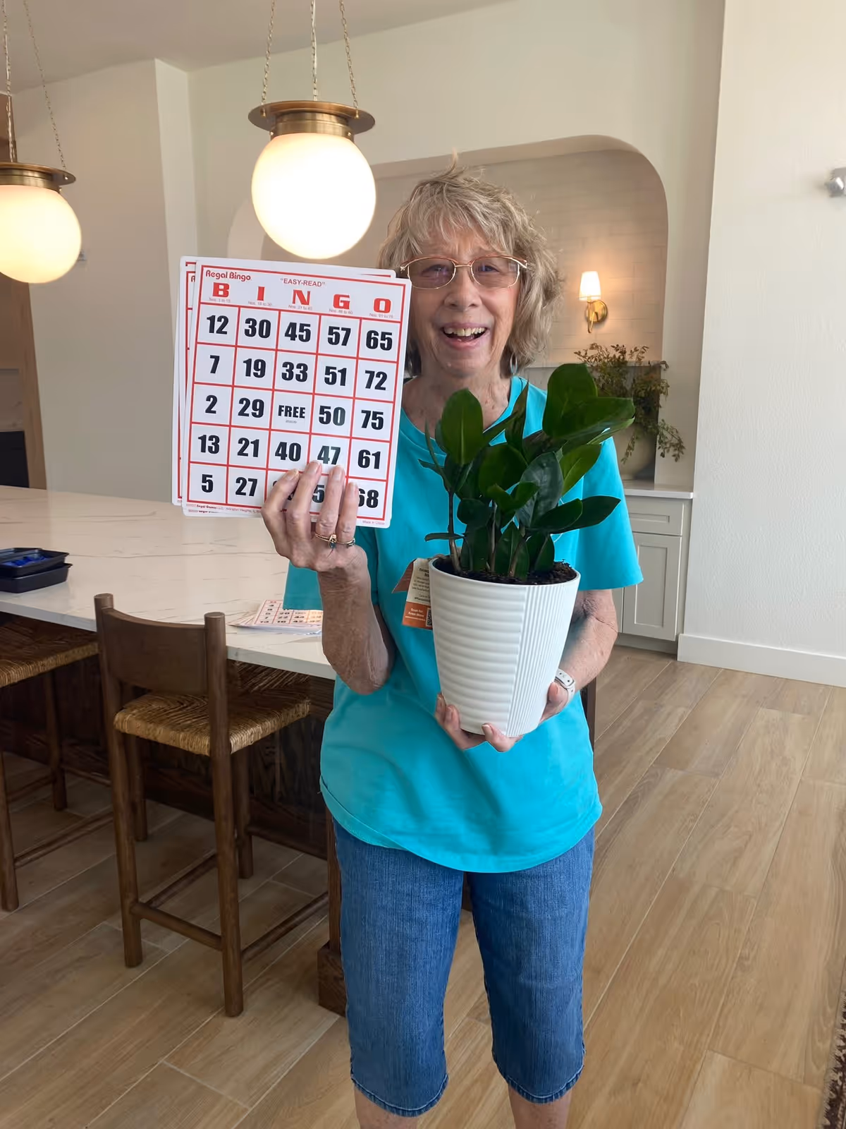An elderly woman wearing glasses and a turquoise shirt stands indoors holding a bingo card in one hand and a potted green plant in the other. Behind her is a kitchen island with wooden chairs and hanging globe pendant lights.