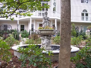 Outdoor garden area with a decorative stone fountain featuring cherub statues in the center, surrounded by greenery and plants. In the background, there is a multi-story white building with balconies and windows.