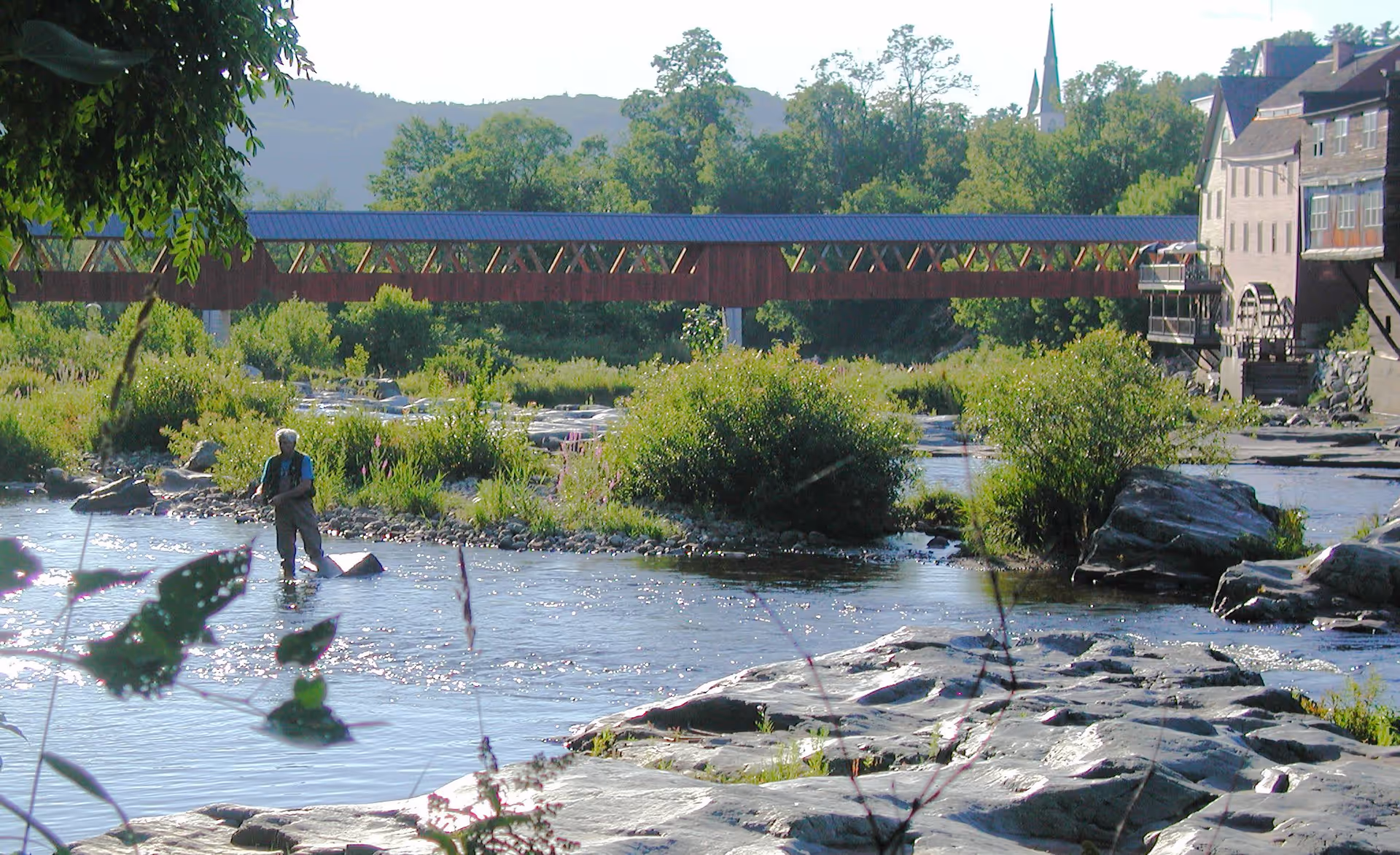 A person standing in a shallow river fishing with a rod, surrounded by rocks and greenery. In the background, there is a covered wooden bridge and a building with a water wheel, with trees and hills further behind.