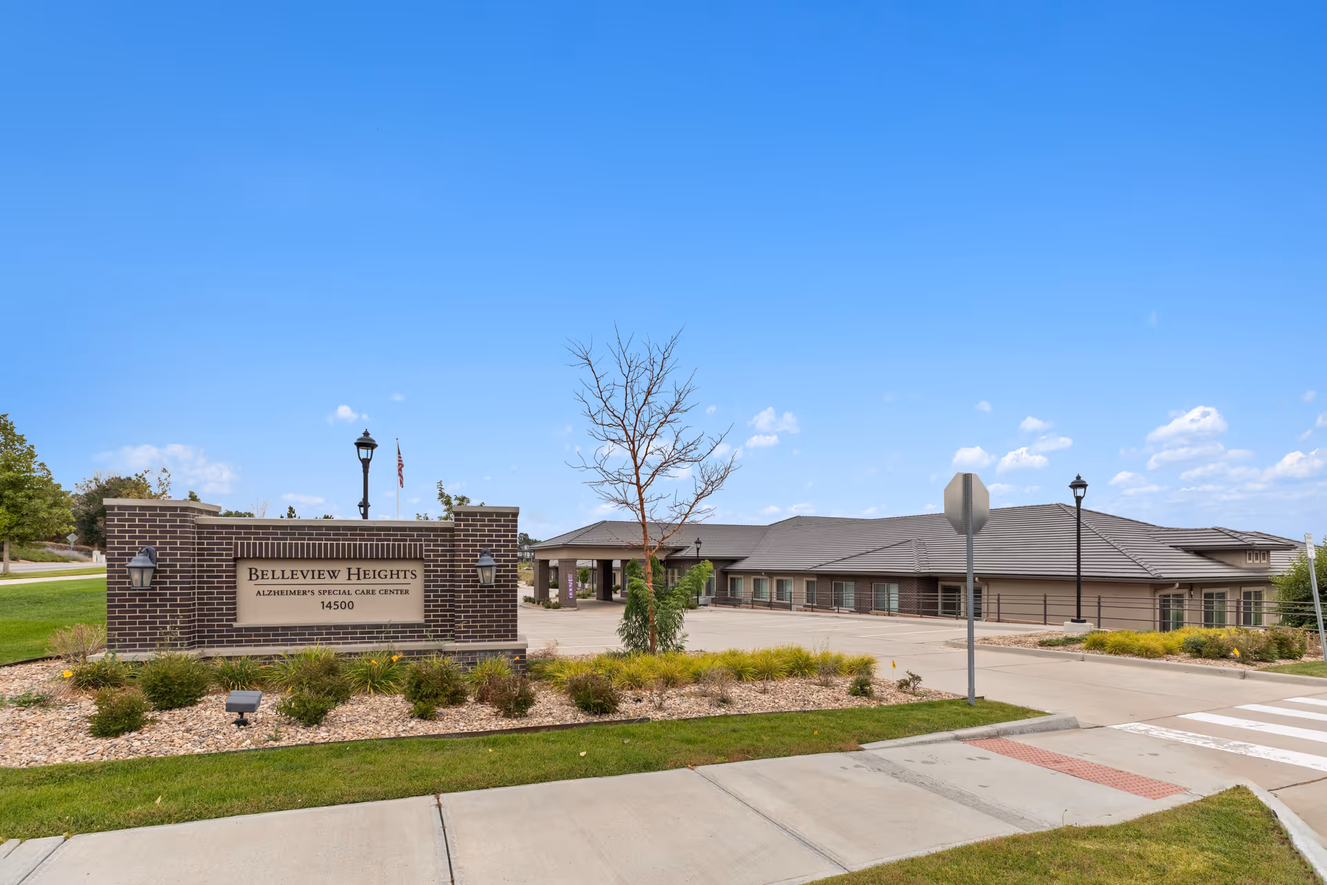 Exterior view of Belleview Heights Alzheimer's Special Care Center building with a brick sign in front displaying the facility name and address. The building is single-story with a gray roof, surrounded by landscaped greenery and a clear blue sky above.