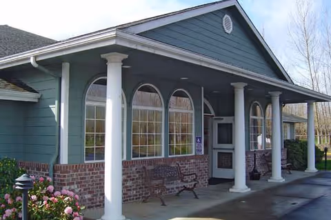 Exterior view of a building with a covered porch supported by white columns. The building has blue siding with brick accents and multiple arched windows. There are benches on the porch and some flowers in the foreground.