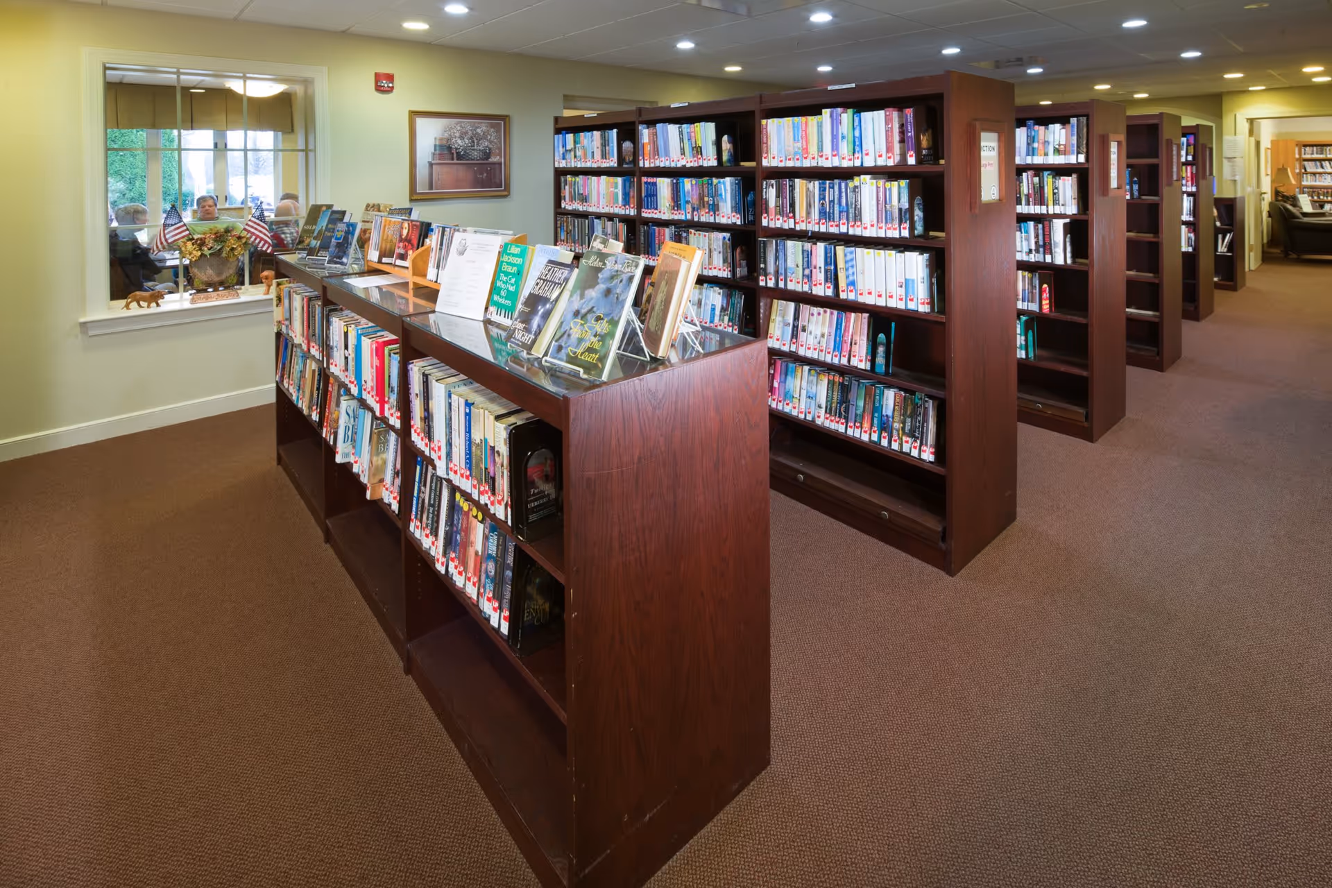 Interior view of a library area in Carroll Lutheran Village with multiple wooden bookshelves filled with books. A display shelf in the foreground showcases several books facing outward. There is a window on the left side with a view into another room where three elderly people are seated. The room is well-lit with ceiling lights and has carpeted flooring.