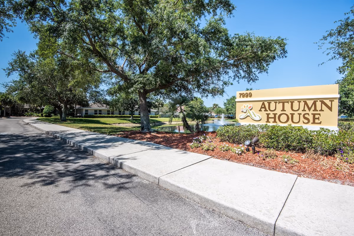 Outdoor view of the Autumn House facility entrance with a large sign displaying the name and address number 7999. The scene includes a paved road, sidewalk, landscaped greenery, trees, and a pond with a fountain in the background under a clear blue sky.