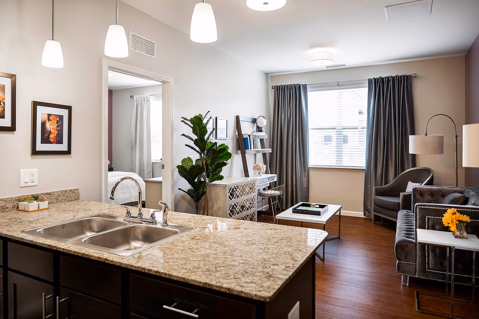Interior view of a senior living facility apartment showing a kitchen island with a double sink and granite countertop in the foreground. Beyond the kitchen is a living area with a gray sofa, armchair, coffee table, floor lamp, and a window with dark curtains. A small desk with a chair and decorative items is against the wall. A doorway leads to a bedroom with a bed and white curtains.
