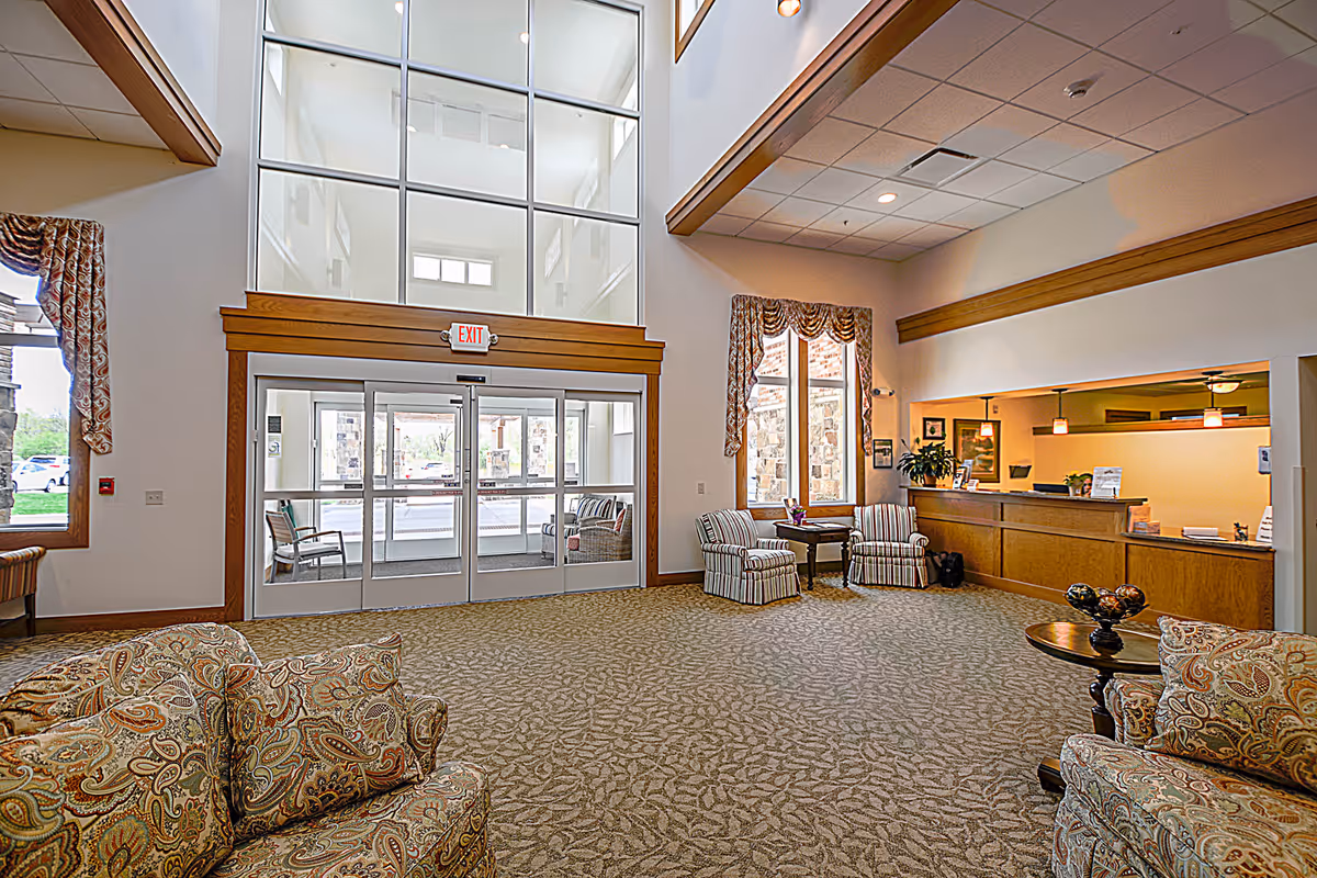 Spacious senior living facility lobby with patterned carpet, floral upholstered armchairs, a wooden reception desk, large windows with curtains, and glass double doors leading outside.