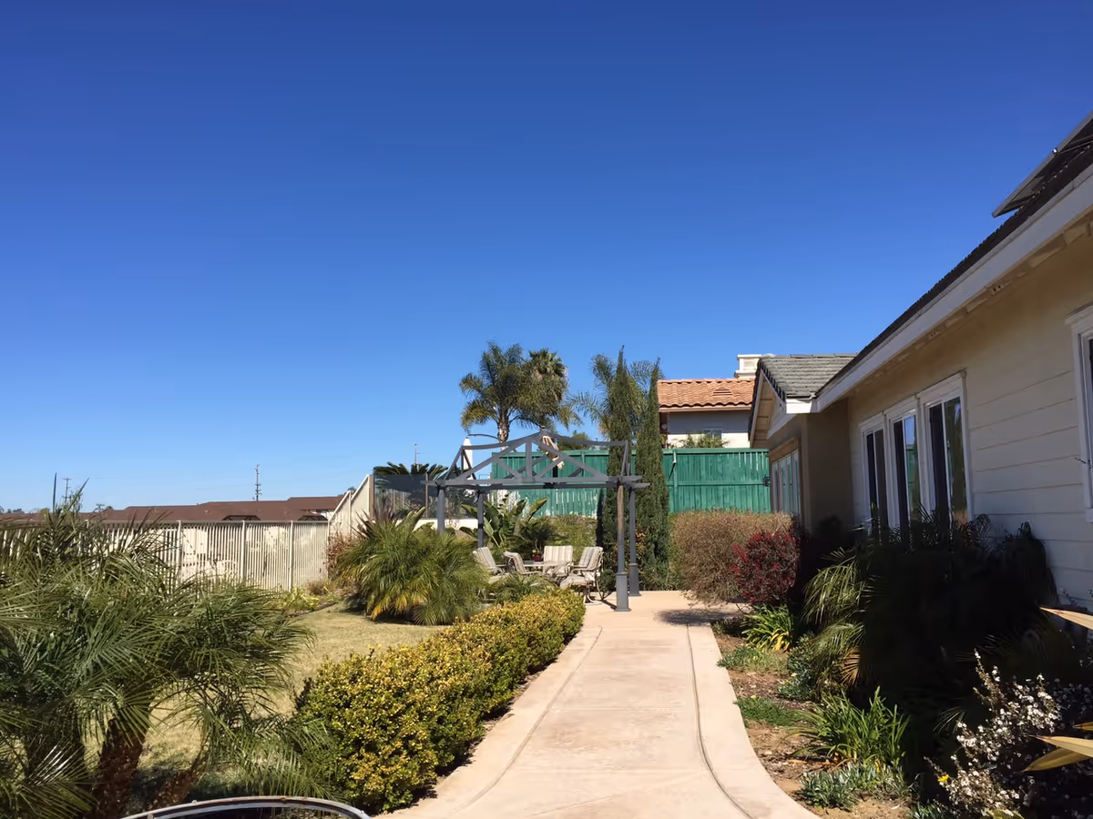 Outdoor pathway leading to a seating area with chairs under a pergola, surrounded by various green plants and bushes. The pathway is adjacent to a beige building with multiple windows. The sky is clear and blue.