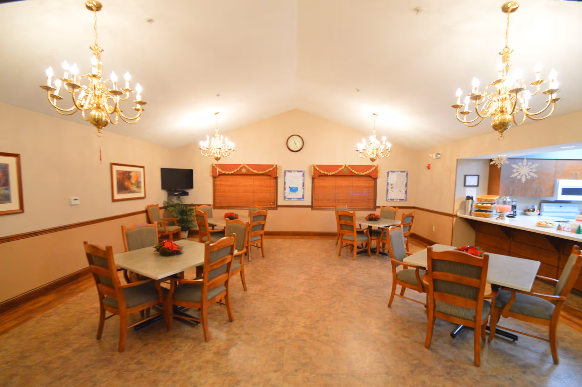 A dining room with several square tables, each surrounded by four wooden chairs with cushioned seats. The room has beige walls with wooden trim, three chandeliers hanging from the ceiling, and two windows with wooden blinds and decorative valances. A clock is mounted on the wall between the windows. There is a small television mounted on the left wall and a kitchen or serving area visible on the right side with a counter, microwave, and refreshments.