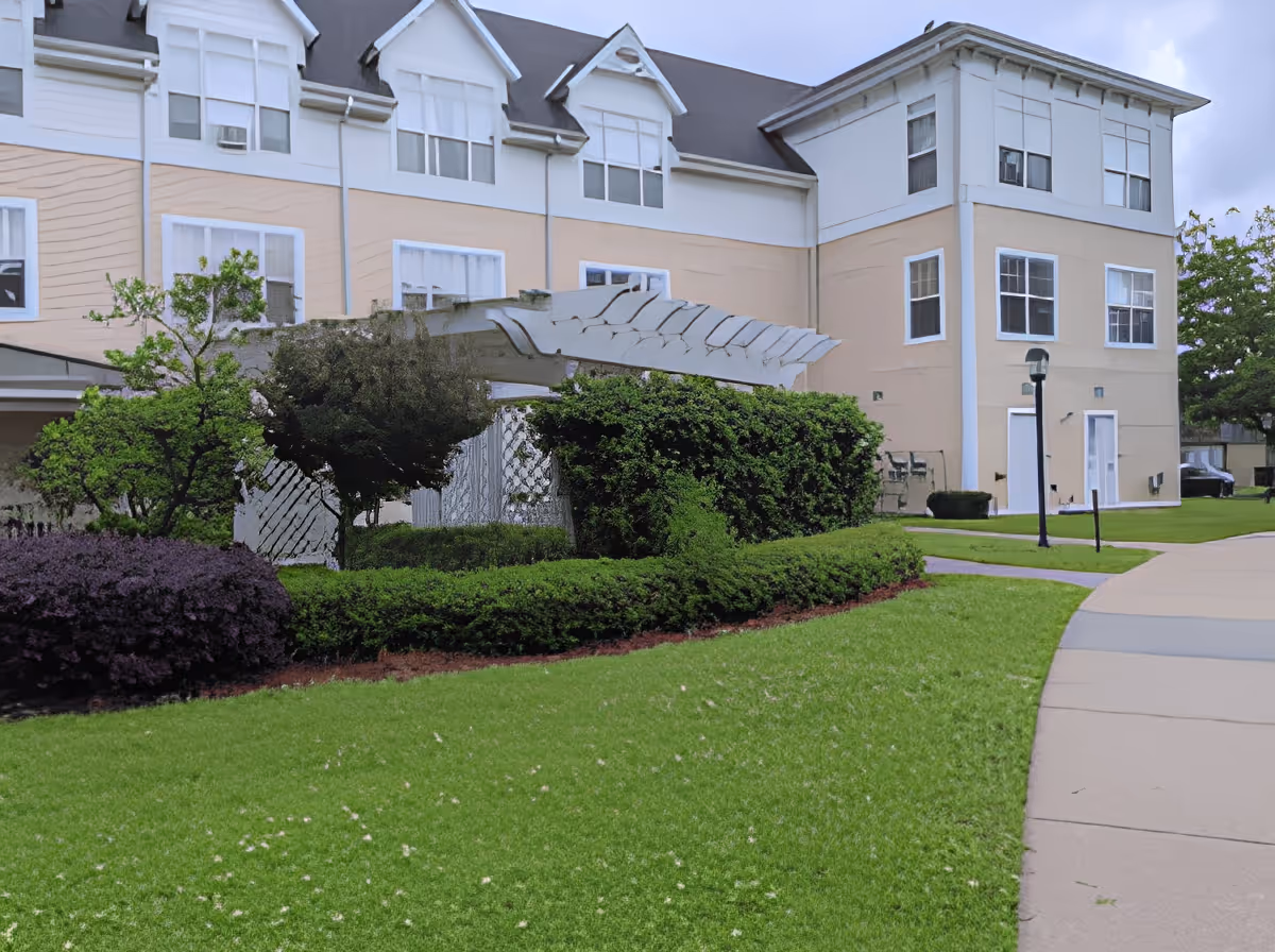 Exterior view of a multi-story senior living facility building with beige and white walls, multiple windows, and a dark roof. In front of the building, there is a well-maintained green lawn, bushes, and a white pergola structure surrounded by greenery. A paved walkway curves along the right side of the image with a streetlamp near the building.