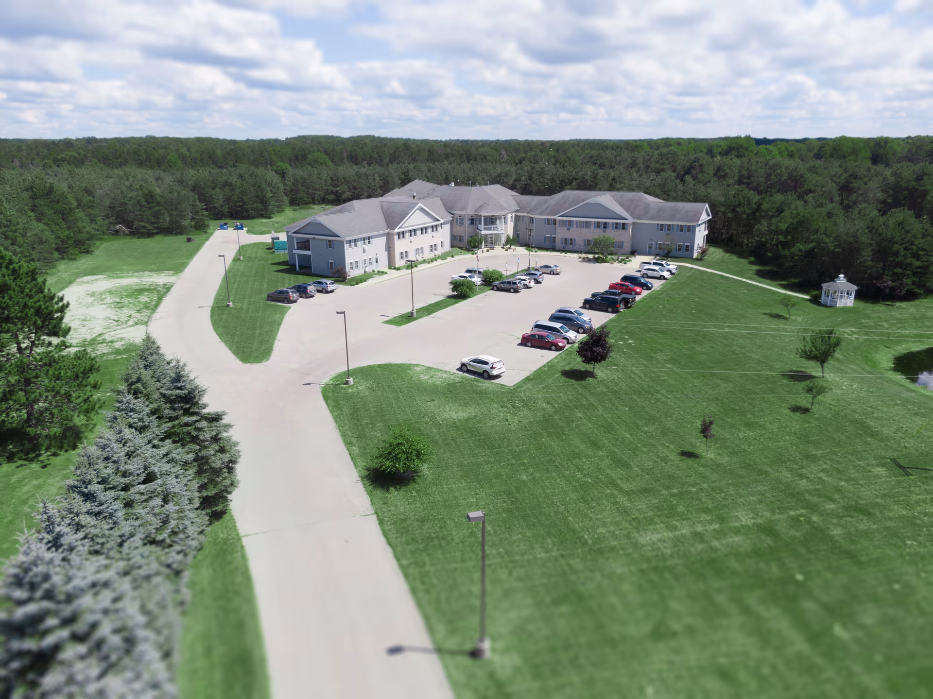 Aerial view of Whispering Pines Senior Living facility showing a large building surrounded by green lawns and trees. There is a parking lot with several cars parked, a paved driveway, and a small white gazebo on the right side. The sky is partly cloudy.